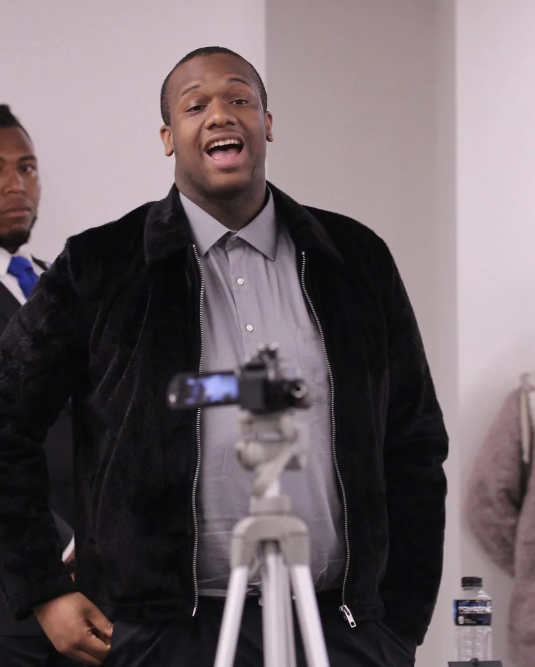 Kenneth, wearing a grey button-down and black jacket, speaks before a student government meeting.